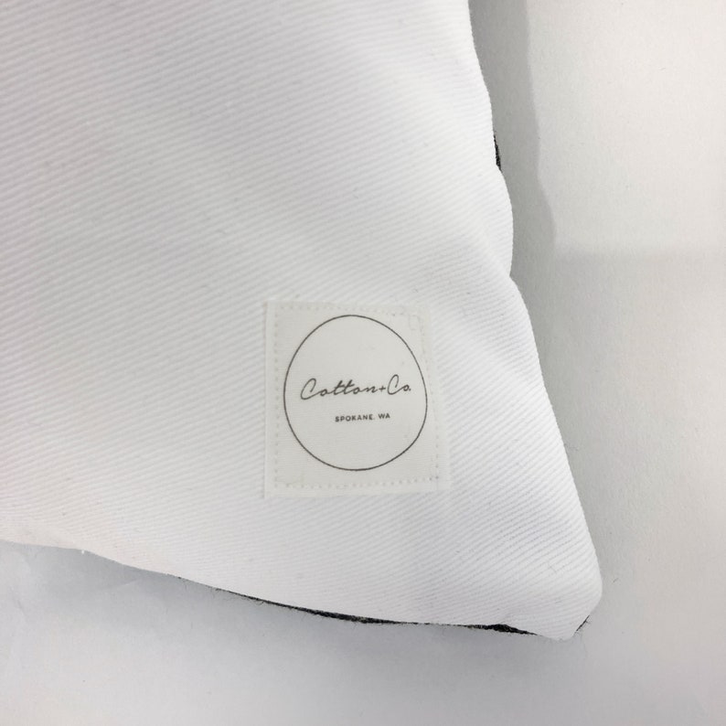 May include: White pillow with a black border and a white label with the text "Cotton+Co. Spokane, WA" in black.