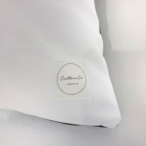 Pu&ograve; includere: Cuscino bianco con bordo nero e un'etichetta bianca con il testo "Cotton+Co. Spokane, WA" in nero.
