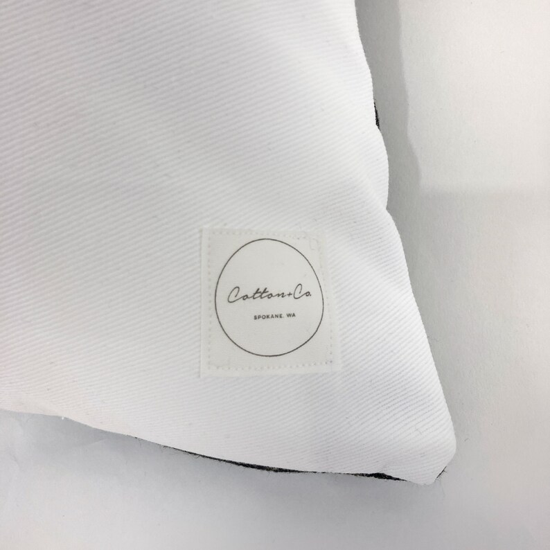 Pu&ograve; includere: Cuscino bianco con bordo nero e un'etichetta bianca con il testo "Cotton+Co. Spokane, WA" in nero.