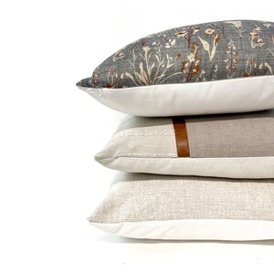 May include: Three decorative throw pillows stacked on top of each other. The top pillow has a floral pattern in shades of gray, white, and orange. The middle pillow is a light gray with a brown leather accent. The bottom pillow is a light beige with a subtle texture.