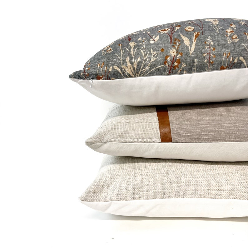 May include: Three decorative throw pillows stacked on top of each other. The top pillow has a floral pattern in shades of gray, white, and orange. The middle pillow is a light gray with a brown leather accent. The bottom pillow is a light beige with a subtle texture.