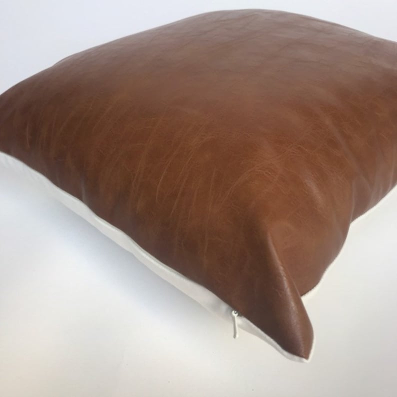 May include: A brown faux leather pillow with a white zipper and white piping around the edges.