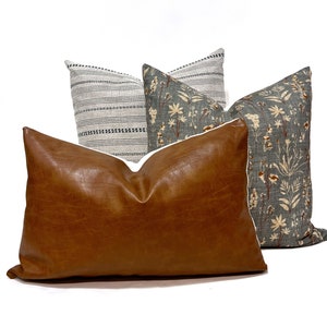 Pu&ograve; includere: Tre cuscini decorativi, un cuscino in similpelle marrone con bordino bianco, un cuscino a righe grigio e bianco con bordino nero e un cuscino floreale grigio con bordino bianco.