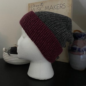 Könnte beinhalten: Handgestrickte Mütze in Grau und Burgunderrot. Die Mütze wird auf einem weißen Schaufensterkopf präsentiert. Der burgunderrote Rand ist hochgekrempelt, und der graue Teil der Mütze ist locker. Im Hintergrund sind ein Schallplattencover und eine Keramikvase zu sehen.