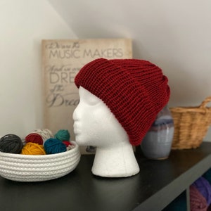Könnte beinhalten: Eine gestrickte, weinrote Beanie-Mütze, die auf einem weißen Styroporkopf präsentiert wird. Die Mütze besteht aus einem Rippenstrickstoff. Im Hintergrund befindet sich eine weiße Schale mit verschiedenen farbigen Garnknäueln und ein Vintage-Druck.