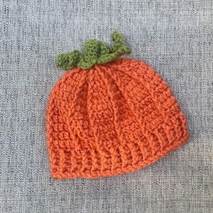 May include: An orange crochet hat with a green stem, resembling a pumpkin. The hat is made of a textured yarn and is likely for a baby or toddler. The hat is a festive accessory for Halloween or fall.