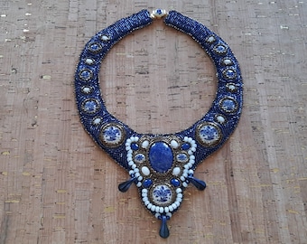 Puede incluir: Un collar de cuentas azul y blanco con un gran colgante de lapislázuli. El colgante está rodeado de cuentas más pequeñas de color blanco y azul y tiene un diseño floral.