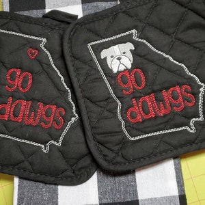 May include: Two black quilted pot holders with white stitching. One features a heart and the words "go dawgs" in red, the other has a bulldog graphic and the same text. A black and white checkered towel is visible.
