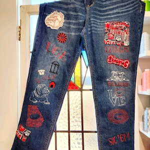 May include: Blue denim jeans with various patches and embroidery. The patches include a bulldog, a state of Georgia outline, and the words "UGA", "Dawgs", "Bulldogs", "Love", "Sic 'Em", and "Go Dawgs".