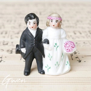 May include: A porcelain figurine of a bride and groom. The bride is wearing a white dress with green floral accents and a pink flower crown. The groom is wearing a black suit and a top hat. The figurine is standing on a sheet of music.