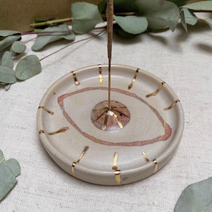 May include: A white ceramic incense holder with a gold eye design and a single incense stick. The holder has a small, raised platform in the centre for the incense stick to rest on.