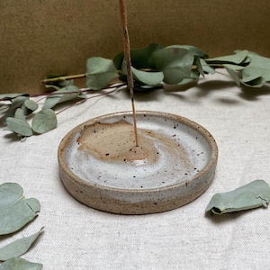 May include: A white ceramic incense holder with a brown speckled glaze. The holder has a small, raised centre for holding incense sticks.