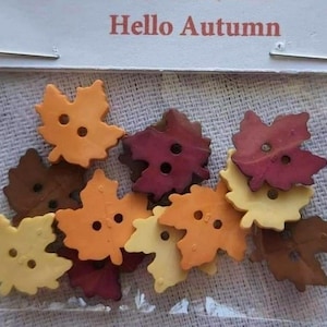 May include: A collection of leaf-shaped buttons in shades of orange, yellow, brown, and burgundy, each featuring two center holes. The buttons are packaged in a clear plastic bag with a white header card displaying the text Hello Autumn.