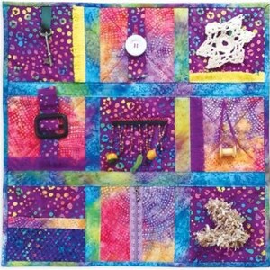 May include: A colorful sensory board with various textures and objects. It features a key, button, buckle, beaded fringe, crochet star, and a spool of thread. The board is primarily purple with blue, yellow, and pink accents, designed for tactile exploration.
