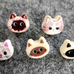 May include: Five small, cream-colored buttons shaped like cat heads. Each button features a unique feline face with distinct markings, including Siamese-style points, tabby stripes, and solid black ears, all with detailed eyes, noses, and whiskers.