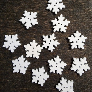 May include: Twelve white wooden snowflake buttons with a raised design. The buttons are arranged in a random pattern on a dark brown surface.
