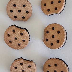 May include: Six brown cookie-shaped embellishments with dark brown chocolate chip details. The cookies have a light brown exterior and a dark brown filling. Some cookies have a bite taken out of them.
