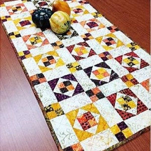 May include: A white table runner with a patchwork design featuring squares of yellow, orange, brown, and purple fabric. The runner is decorated with three small gourds.