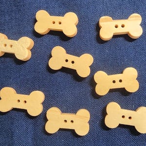 May include: Eight wooden bone-shaped buttons in a light brown color. The buttons have two holes each.