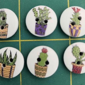 May include: Six round, white wooden buttons with cactus designs in various pots. The cacti are green with red, purple, and yellow pots. Each button has two black holes for sewing.