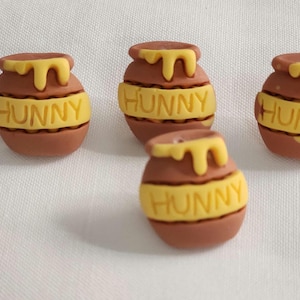 May include: Four brown clay pots with yellow honey drips at the top and a yellow band around the center featuring the word HUNNY embossed in capital letters. The pots are arranged in a cluster, each showing the same design and color scheme.