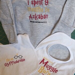May include: A gray long-sleeve onesie with the text "I spent 9 months in Azkaban" and a black wand underneath. Two white bibs with embroidered text. One bib says "I'm so Gryffindorable" with a pair of glasses and a lightning bolt. The other bib says "Muggle Born" with a wand.