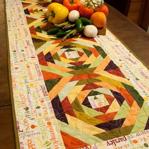 May include: A quilted table runner with a geometric pattern in shades of orange, green, and brown. The runner features a white background with text printed in black, listing various vegetables and herbs. The runner is decorated with fresh vegetables, including red onions, tomatoes, peppers, asparagus, and cauliflower.