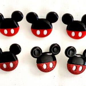 May include: Six small buttons shaped like the head of a cartoon mouse. The top half is glossy black, and the bottom half is red with two white oval accents. Three buttons have a smooth red finish, while the other three have a glittery red finish.