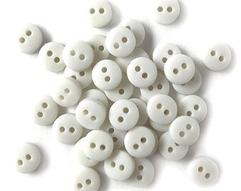 May include: A collection of small, round, off-white buttons with two holes each. The buttons are scattered across a white surface, creating a simple, clean aesthetic. These buttons are ideal for sewing and craft projects.