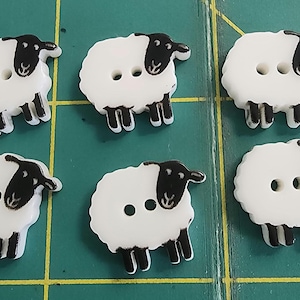 May include: Six sheep-shaped buttons with white bodies and black heads and legs. Each button has two holes for sewing. The buttons are arranged on a green cutting mat.