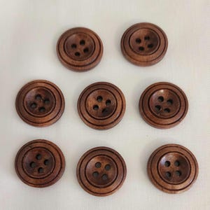 May include: A collection of eight round, wooden buttons with a dark brown finish. Each button features four evenly spaced holes and concentric circle detailing around the center. The buttons are arranged on a white surface.