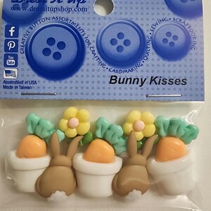 May include: A package of "Bunny Kisses" embellishments for crafting. The package includes decorative buttons and plastic bunny, carrot, and flower shapes in pastel colors. The package text reads "Dress It Up" and "Creative Button Assortments for Crafting".