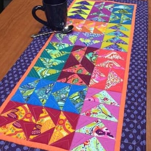 May include: A colorful patchwork table runner with a geometric pattern of triangles in shades of blue, green, orange, red, purple, and yellow. The runner has a purple border and an orange trim.