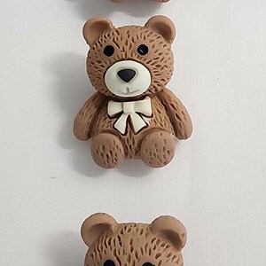 May include: Three brown teddy bear figurines with white bow ties and black eyes are arranged vertically against a white background. The bears have a classic design, with rounded bodies and small ears.