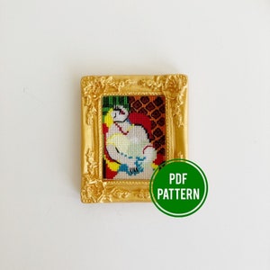 May include: A small, framed cross-stitch artwork featuring a figure in a white dress, with yellow and blue accents, set against a red and brown patterned background. The frame is ornate, gold-colored, and the image includes a green circle with the text "PDF PATTERN".