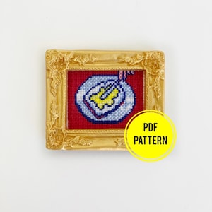 May include: A gold-framed picture with a red background and a cross-stitch design of a plate with a yellow and white cake inside. The text "PDF PATTERN" is on a yellow circle below the picture.