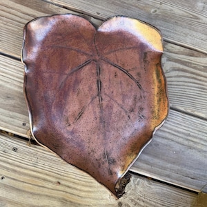 May include: A leaf-shaped ceramic tray featuring a metallic copper and bronze glaze with a dark, mottled finish. The surface displays embossed vein details that mimic the natural structure of a large, heart-shaped foliage leaf.