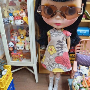 May include: A doll with black hair wearing heart-shaped brown sunglasses and a beige sleeveless dress featuring a floral pattern in yellow, pink, and grey. The doll is wearing white lace-up boots and stands in a room with a glass display cabinet.