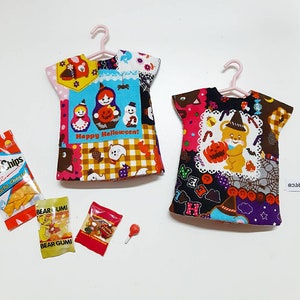 May include: Two miniature doll shirts on pink hangers, one featuring a matryoshka doll and pumpkin print with the text Happy Halloween, the other with a bear and pumpkin print. Included are miniature snack packets labeled Hello Chips and Bear Gumi.