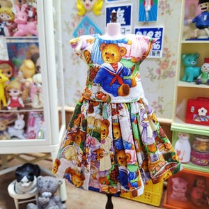 May include: A miniature dress with a teddy bear print. The dress features a white bodice with a teddy bear wearing a sailor outfit. The skirt is a colorful mix of teddy bears and other illustrations. The dress is displayed on a black stand.
