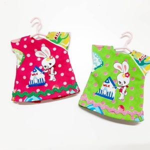 May include: Two doll dresses with a polka dot pattern and a bunny rabbit design. One dress is pink with green trim and the other is green with pink trim. Both dresses have the word "MILK" on them.