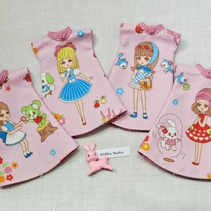 May include: Four pink doll dresses with cartoon illustrations of girls, animals, and floral patterns. The dresses have a round collar with a polka dot design. A small pink bunny figurine is in the foreground.