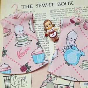 May include: Two pink Kewpie doll dresses with a repeating pattern of cupcakes, roses, and Kewpie dolls. A small Kewpie doll is pinned between the dresses. The dresses are hanging on pink hangers. The text "Kewpie" is printed on the dresses.