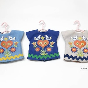 May include: Three miniature doll dresses with embroidered floral designs and birds. The dresses are hanging on hangers and are in blue, white, and beige.