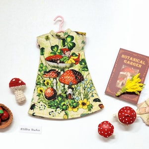 Puede incluir: Un vestido verde y blanco con estampado de setas y hadas. El vestido tiene cuello alto y falda fruncida. El vestido está colgado de una percha rosa. El vestido está rodeado de pequeñas decoraciones de setas y manzanas.