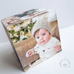 Op de afbeelding: Een fotokubus met een foto van een baby die een witte gebreide muts en trui draagt met een berenontwerp. De baby zit voor een kerstboom.