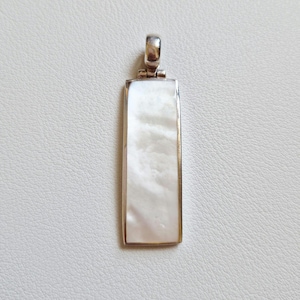 May include: A rectangular pendant with a pearlescent white center and a silver-colored metal frame. The pendant has a loop at the top for a chain. The white center has a subtle, swirling pattern.