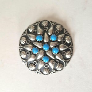 Puede incluir: Broche redondo de metal plateado con diseño floral. El broche presenta siete piedras redondas de color azul brillante en el centro y alrededor del centro. El borde exterior tiene un patrón texturizado detallado.