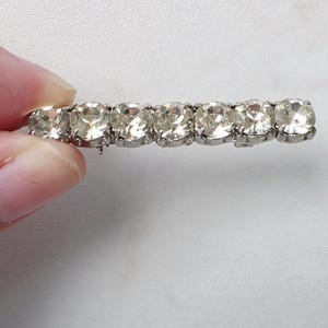 Può includere: Fermaglio per capelli argentato con sei strass ovali trasparenti. Gli strass sono disposti in fila, creando un effetto scintillante. Il fermaglio è tenuto da una mano su sfondo bianco.