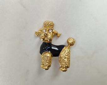 May include: A gold-toned poodle brooch with a black body and sparkling accents. The poodle has a textured coat and is posed in a standing position. The brooch is a vintage-style accessory.